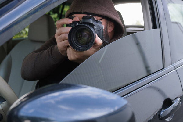 A man in a hood and sunglasses sits in a car and directs the lens of a professional chamber out through an open window. Hidden observation, surveillance or work of a private detective.