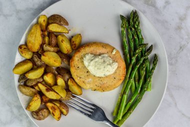 Sotelenmiş kuşkonmaz ve patates dilimleriyle servis edilen tarter soslu somon burger üstü.,