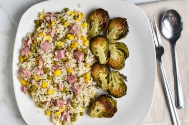 Pirinç kabuğu dilimlenmiş konserve jambon, mısır ve bezelye ile kızartılmış brussel lahanayla servis edilir.,