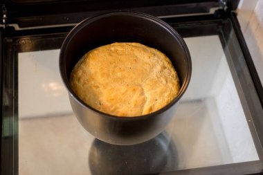 Teflon kaplı tavada ev yapımı ekmek elektrikli fırının açık cam kapısında duruyor. Yüzeyde altın kabuk ve tohumlar. Taze pişmiş somun, çekici bir doku sergiliyor..