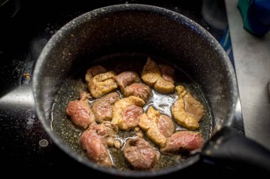 Tavada kızarmış ördek eti. Parçalar cızırdıyor. Yağ köpürüyor. Siyah yüzey görünüyor. Meyve suları parlıyor. Kenarları gevrek. Yemek pişirme devam ediyor. Zengin aromalı mutfak. Ilık ışıklandırma. Ev hazırlığı.