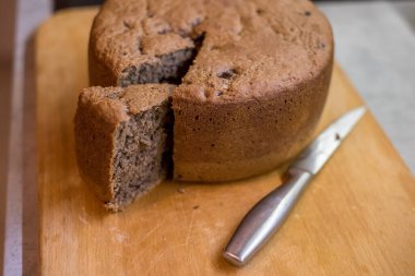Tahtaya yerleştirilmiş yuvarlak pasta yanında metal bıçakla, kalın bir dilim kesilmiş. Yüzey çatlaklarla ve pişmiş dokularda görülebilen koyu dolgularla sıkı görünüyor..