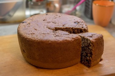 Taze pişmiş kek tahtanın üzerinde duruyor, bir dilim kesilmiş. Arka plan bulanık, mutfak malzemelerini incelikle gösteriyor. Sahne sıcaklık, doku, detay, ışık, ruh hali, lezzet yakalar.