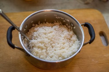 Pişmiş pirinç tencerenin içinde nazikçe yerleştirilmiş kaşıkla dinleniyor. Buhar yavaşça akıyor. Basit mutfak atmosferi sıcaklık, tat, doku, durgunluk, denge, ışık, bakım, konfor öneriyor.