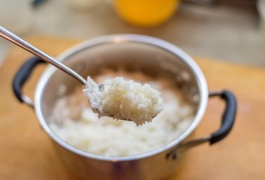 Kaşık, pilavın bir kısmını kameraya yakın tutuyor, tek tek taneler görünüyor. Arka planda tahtanın üzerine yerleştirilmiş bulanık tencere görünüyor. Parlak metal yüzeyden yana uzanan siyah saplar..