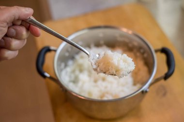 Tahtaya yerleştirilmiş metal tencerenin üzerinde pişmiş pirinç içeren el kaldırma kaşıkları. Buhar hafifçe görünür. Mutfak ortamı bulanık. Sapları çatal bıçak kullanımını açıkça gösteriyor.