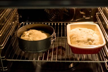 Fırın rafında yükselen hamurlu seramik ve metal tavalar var. İçeride ışık parlıyor. Ekmek eşit olarak pişirmeye başlar. Sıcak atmosfer, modern mutfak ortamında yemek pişirme sürecini çevrelemektedir..