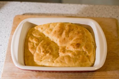 Taze pişmiş somun, güverteye yerleştirilmiş seramik tabağın içinde duruyor. Ekmek yüzeyi altın gibi görünüyor ve biraz düzensiz. Kenarları yavaşça yükseliyor. Arka plan yumuşakça harmanlanır, rahat mutfak havasını arttırır..