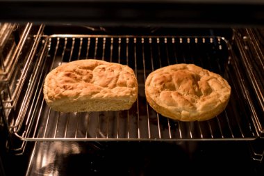 İki yeni pişmiş ev yapımı ekmek, metal fırın rafında duruyor, farklı şekiller, sıcak ışık altında parlayan altın kabuklar, kabarık doku görünür, pişirme süreci neredeyse bitti, mutfak atmosferi rahat..