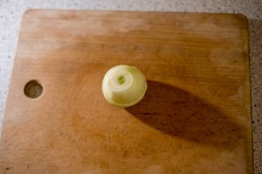 Soyulmuş soğan, tahta tahtanın üzerinde sessizce yatar ve pürüzsüz yüzeyde uzun bir gölge bırakır. Işık ampulün üzerine hafifçe düşer, ince çizgiler ve dokular dilimlenmeyi ya da doğramayı beklerken vurgular..