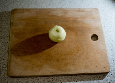 Soyulmuş soğan, tahta tahtanın üzerinde sessizce yatar ve pürüzsüz yüzeyde uzun bir gölge bırakır. Işık ampulün üzerine hafifçe düşer, ince çizgiler ve dokular dilimlenmeyi ya da doğramayı beklerken vurgular..