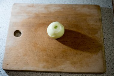 Soyulmuş soğan, tahta tahtanın üzerinde sessizce yatar ve pürüzsüz yüzeyde uzun bir gölge bırakır. Işık ampulün üzerine hafifçe düşer, ince çizgiler ve dokular dilimlenmeyi ya da doğramayı beklerken vurgular..