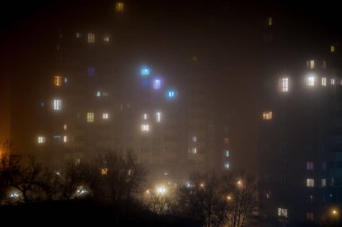 Sisli gece manzarası parlayan yüksek binaların camlarını ortaya çıkarıyor. Ön plandaki ağaçlar sisli ambiyansla harmanlanır. Işıklar birden fazla rengi yansıtıyor, kentsel sükuneti ve derinliği gösteriyor.