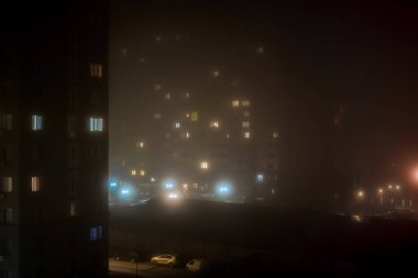 Sisli gece manzarası parlayan yüksek binaların camlarını ortaya çıkarıyor. Ön plandaki ağaçlar sisli ambiyansla harmanlanır. Işıklar birden fazla rengi yansıtıyor, kentsel sükuneti ve derinliği gösteriyor.