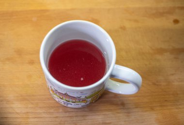 Kupa, iç boşluğu dolduran kırmızı komposto içeriyor. Konteynır ahşap masa yüzeyinin üzerinde duruyor. Mutfak alanı yemek hazırlama anlamına geliyor. Sapı sağdan dışa doğru uzanıyor. Sıvı parlak parlaklığı gösterir.