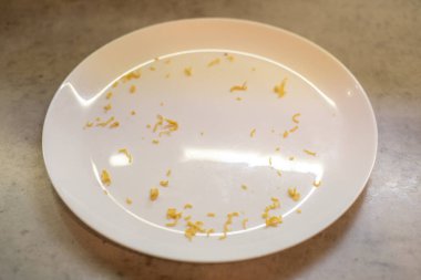 Beyaz plaka etrafa saçılmış vermicelli kalıntılarını barındırıyor. Yüzeye yayılmış küçük kırıntılar. Mutfak tezgahı altında. Düzenleme, önceki yemek tüketimini, bulaşığın temizlenmemiş kalmasını yansıtıyor