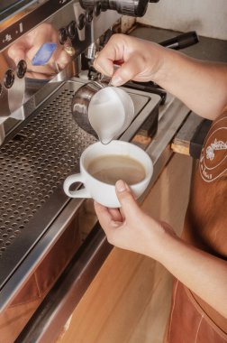 Kafkasyalı bir kadın espresso, latte, latte içinde çikolata sanatı yapıyor. Kafede bir bardak sütle figür yapmak için elinde kap tutuyor. Otomatik makine. Taze öğütülmüş kahve. Kapalı açı. İç.