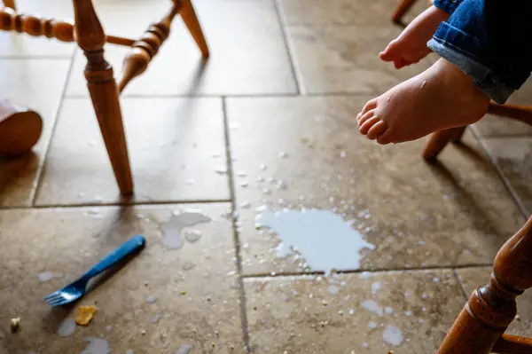 toddler foot suspended above spilled milk, crumbs, and a plastic fork on a brown tile floor . High quality photo