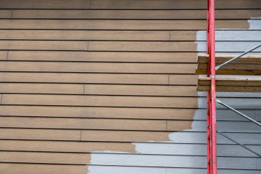 İnşaat iskelesi evin yenilenmesini destekliyor çünkü bir işçi dış cepheye taze boya sürüyor. Bir taraf yeni ve temiz bir palto, diğer taraf ise bitmemiş. Güneş ışığı işin altını çizer.