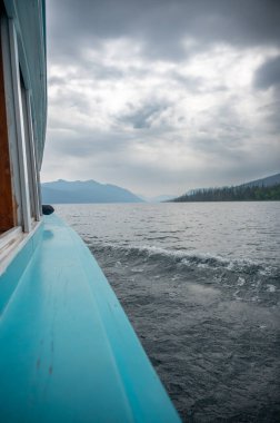 Huzurlu bir gölün sakin sularında süzülen bir tekne ve yükselen sisli dağlar. Gökyüzü bulutlu, doğa ve macerayı birbirine bağlayan sakin bir atmosfer yaratıyor..
