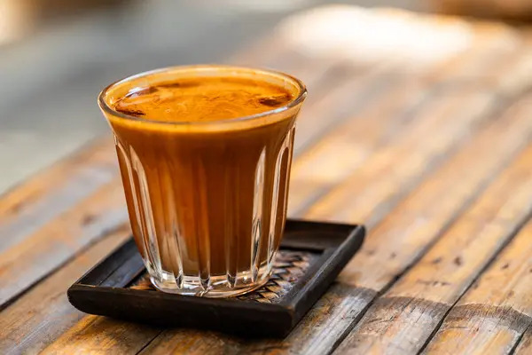 A delicious cup of coffee sits on a bamboo table. The coffee is dark and rich, with a smooth, creamy head. The cup is made of clear glass, and the table is made of light brown bamboo. The background