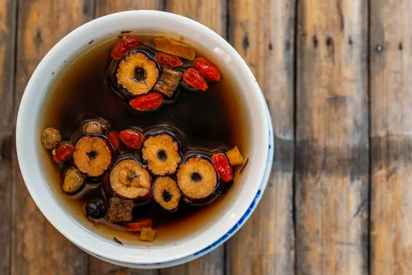 A close-up image of a cup of traditional Chinese herbal tea on a wooden table. The tea is made from a variety of herbs, including dried red dates, goji berries, and chrysanthemum flowers. The tea is