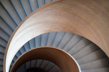 Yukarıdan modern spiral merdiven manzarası. Merdiven pürüzsüz, açık renkli ahşapla yapılmış, parlak ve zarif bir görünüm yaratıyor. Adımlar betondan yapılmış, bir zıtlık ekliyor.