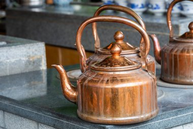 Üç bakır çaydanlık, sıcak renkleri yansıtan bir taş tezgahta oturur. Sahne, yerel bir çay evinin hareketli ruhunu yakalıyor..