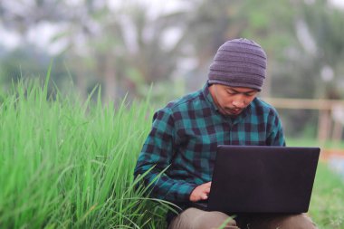 Endonezyalı bereli ve yeşil flanel gömlekli genç adam Rice Fields 'ın ortasında uzaktan çalışıyor..
