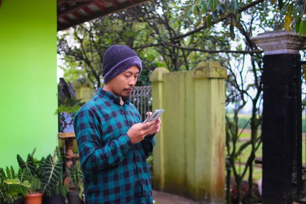 an Asian young man with beanie hat and green flannel shirt is playing with his phone in the backyard.