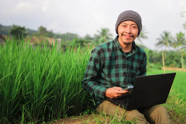 Indonesian young man with beanie hat and green flannel shirt working Remotely at the middle of Rice Fields.