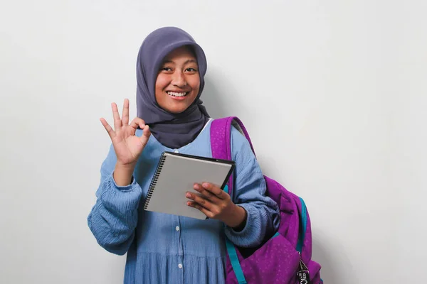 stock image Clever young Asian girl student wearing hijab and backpack, showing OK gesture while holding a pen and book isolated on white background.