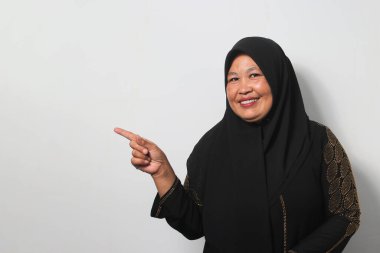Heyecanlı Orta yaşlı, siyah tesettür ve abaya giyen Asyalı kadınlar, beyaz arka planda izole edilmiş kameraya bakarken fotokopi alanının solunu işaret ediyorlar..