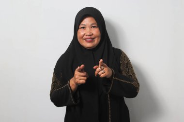 Heyecanlı Orta yaşlı Asyalı kadınlar siyah başörtüsü ve abaya takıyor, beyaz arka planda izole edilmiş kameraya bakarken kamerayı işaret ediyorlar..