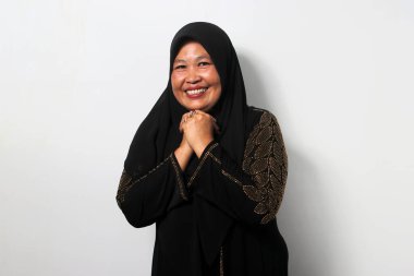 Heyecanlı Orta yaşlı Asyalı kadınlar siyah başörtüsü ve abaya takıyor, yumruğunu çenesine dayayıp beyaz arka planda izole edilmiş bir şey umuyor..