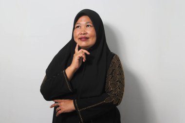 Orta yaşlı Asyalı kadınlar siyah başörtüsü takıyor ve abaya, şüpheye konsantre oluyorlar, parmakları çenesinde ve yukarı bakıp beyaz arka planda izole edilmiş merak ediyorlar..