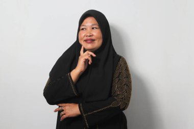 Orta yaşlı Asyalı kadınlar siyah başörtüsü takıyor ve abaya, şüpheye konsantre oluyorlar, parmakları çenesinde ve yukarı bakıp beyaz arka planda izole edilmiş merak ediyorlar..