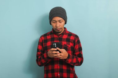 Mutsuz Asyalı adam, bereli ve flanel gömlekli mavi arka planda duran telefonu tutuyor.