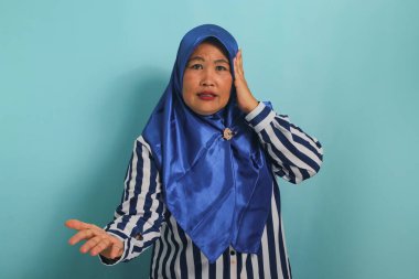 Mavi tesettürlü, çizgili tişörtlü, sinirli, orta yaşlı bir Asyalı kadın mavi bir arka plana karşı durarak el kol hareketi yapıyor.