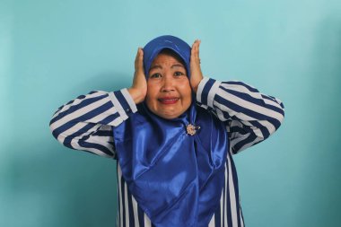 Mavi tesettürlü ve çizgili gömlekli kızgın Asyalı kadın kulaklarını kapatıyor, mavi arka planda gürültüyü durdurmak için el kol hareketi yapıyor.