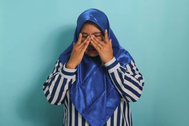 Mavi tesettürlü ve çizgili tişörtlü orta yaşlı bir Asyalı kadın, mavi arka planda izole edilmiş konjonktivit nedeniyle kaşıntı hissettikten sonra gözlerini ovmak için elini kullanır.