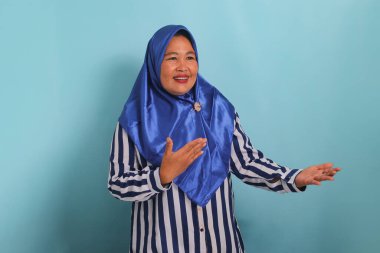 Orta yaşlı bir Asyalı kadın, mavi başörtüsü ve çizgili bir gömlek giyiyor, ellerini yana doğru uzatıyor, birini davet ediyor, mavi arka planda dururken
