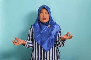 Mavi tesettürlü ve çizgili tişörtlü, rahat, orta yaşlı bir Asyalı kadın, gözleri kapalı meditasyon yapıyor, yoga yapıyor ve ellerini Mudra jesti ile tutuyor. Mavi bir arkaplanda izole edilmiş