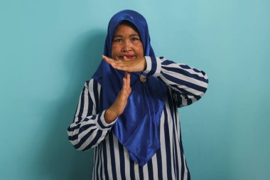 Orta yaşlı, mavi tesettürlü, çizgili tişörtlü, asyalı bir kadın mavi arka planda dikilirken el kol hareketi yapıyor..