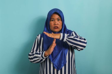 Orta yaşlı, mavi tesettürlü, çizgili tişörtlü, asyalı bir kadın mavi arka planda dikilirken el kol hareketi yapıyor..