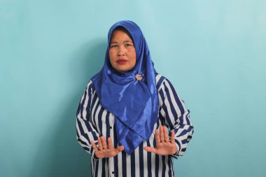 Mavi tesettürlü ve çizgili tişörtlü ciddi bir orta yaşlı Asyalı kadın mavi arka planda dururken dur ya da reddet hareketi gösterir.