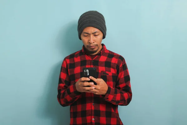 Unhappy Asian man with beanie hat and red plaid flannel shirt holding phone standing over blue background
