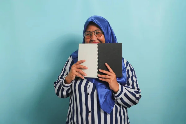 Excited Asian middle-aged woman, wearing a blue hijab, eye glasses and a striped shirt, hides her face behind an open book while looking away, standing against a blue background.