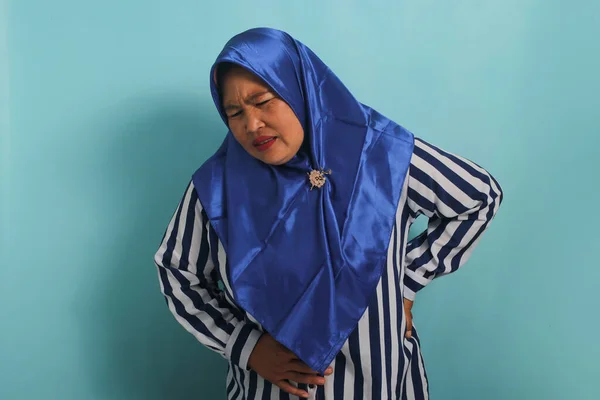 ヒジャーブのアジア系の中年女性の肖像画は 青い背景を背景に立ちながら 彼女が腕で胃をつかんで曲げ 月経痛の痛みを表現していることを示しています — ストック写真