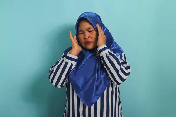 青のヒジャーブとストライプのシャツを着た中年のアジア人女性の肖像画は 青い背景を背景に 片頭痛 または病気に苦しんでいることを示しています — ストック写真